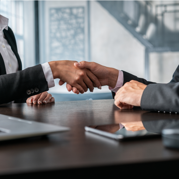 Two businesspeople shaking hands across a table with laptops and tablets in an office setting