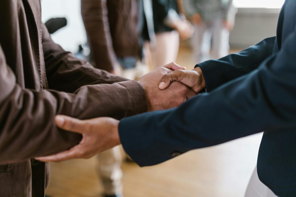 Two people shaking hands and closing the private money lending deal. The background is slightly blurred, showing other individuals standing in the distance, suggesting a formal or professional environment.
